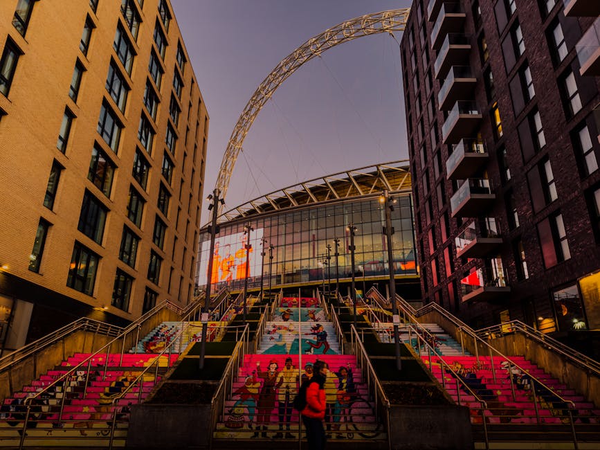 A view of Wembley Stadium's interior showing the empty seating area with bright red upholstered seats arranged in curved rows around the field. The field is covered in well-maintained, green grass wit